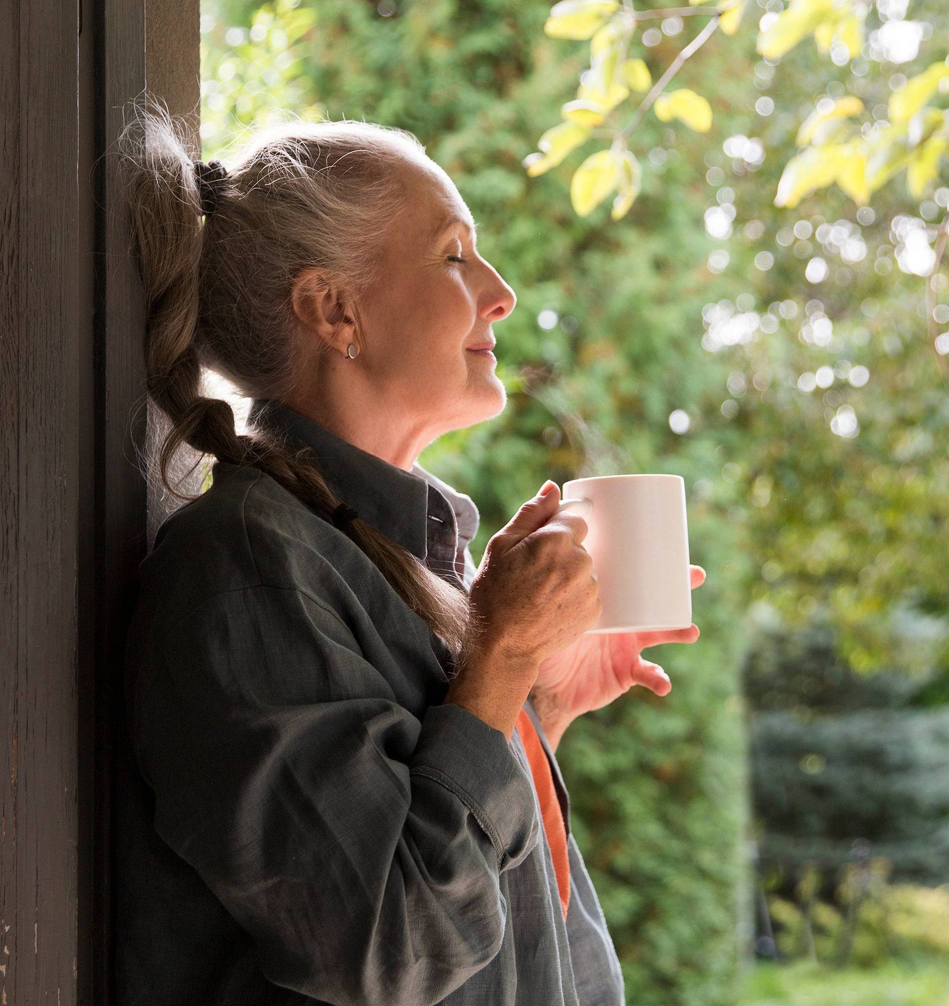 Frau hält eine Tasse am Fenster und genießt einen ruhigen Moment mit Blick ins Grüne.