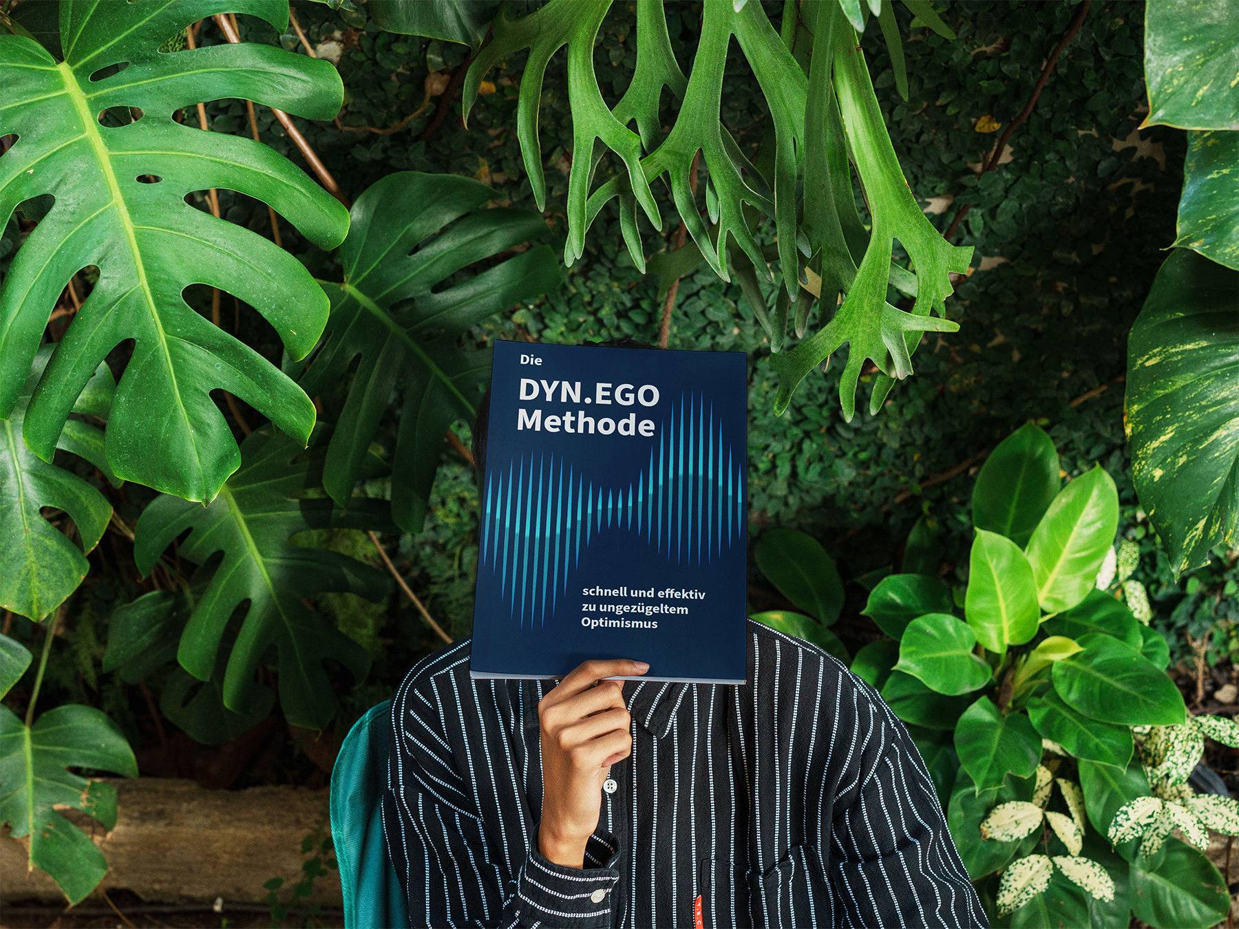 Person hält das DYN.EGO Methodenbuch vor das Gesicht, umgeben von tropischen Grünpflanzen.